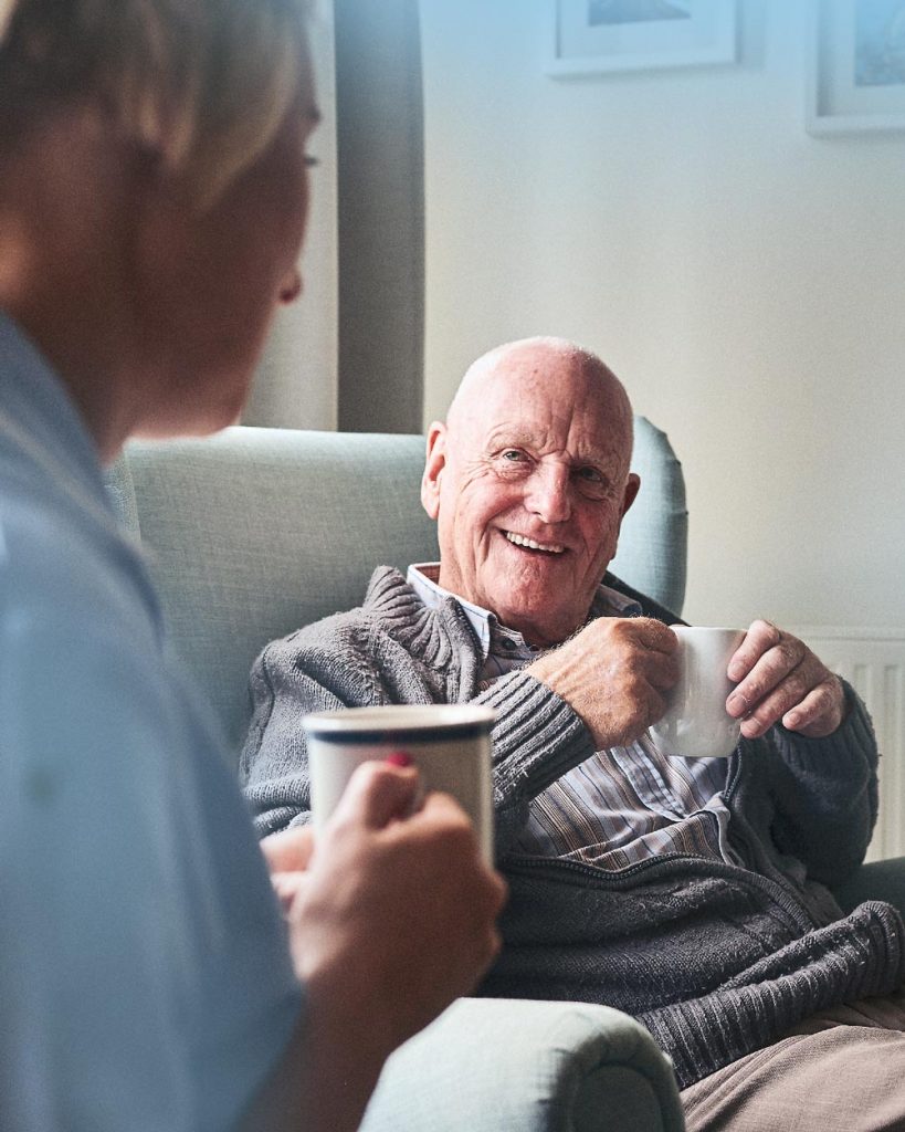 Seniorenbetreuung: Betreuungskraft sitzt zusammen mit einem Senioren im Wohnzimmer bei einer Tasse Tee und unterhalten sich. Der Rentner lächelt die Hygge Betreuung Alltagshilfe an.