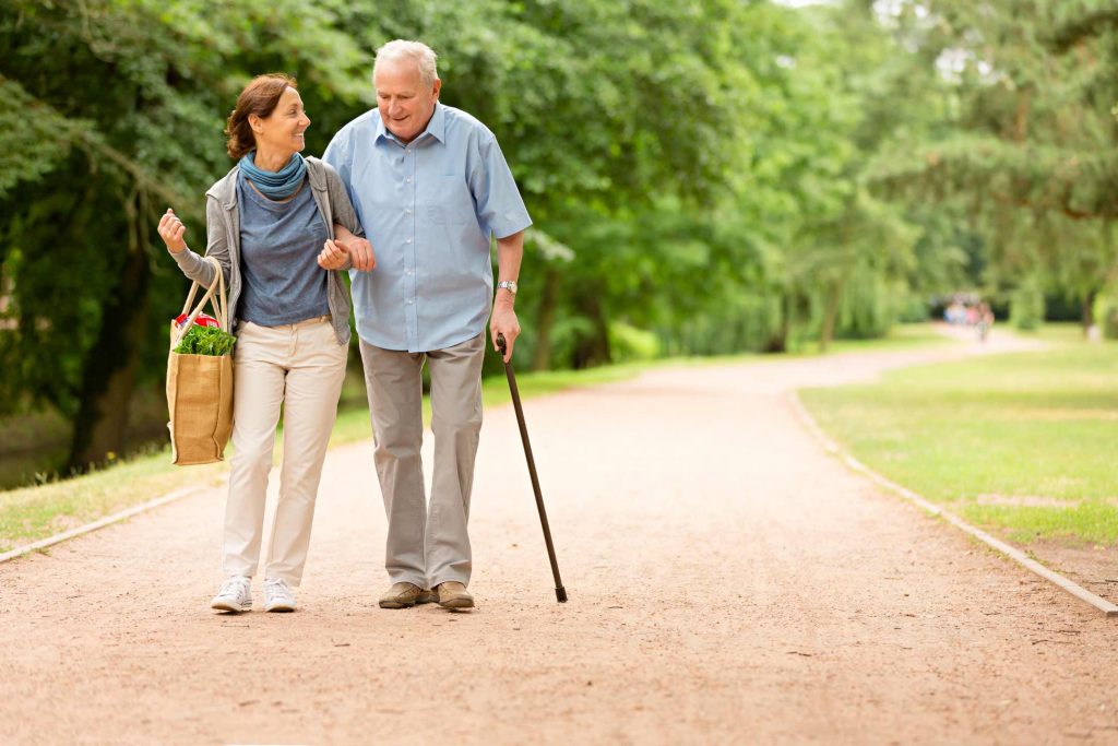 Betreuung und Pflege eines Senioren: Betreuungskraft spaziert mit Senioren durch den Park und trägt einen Korb mit Einkäufen, während sie den Mann stützt