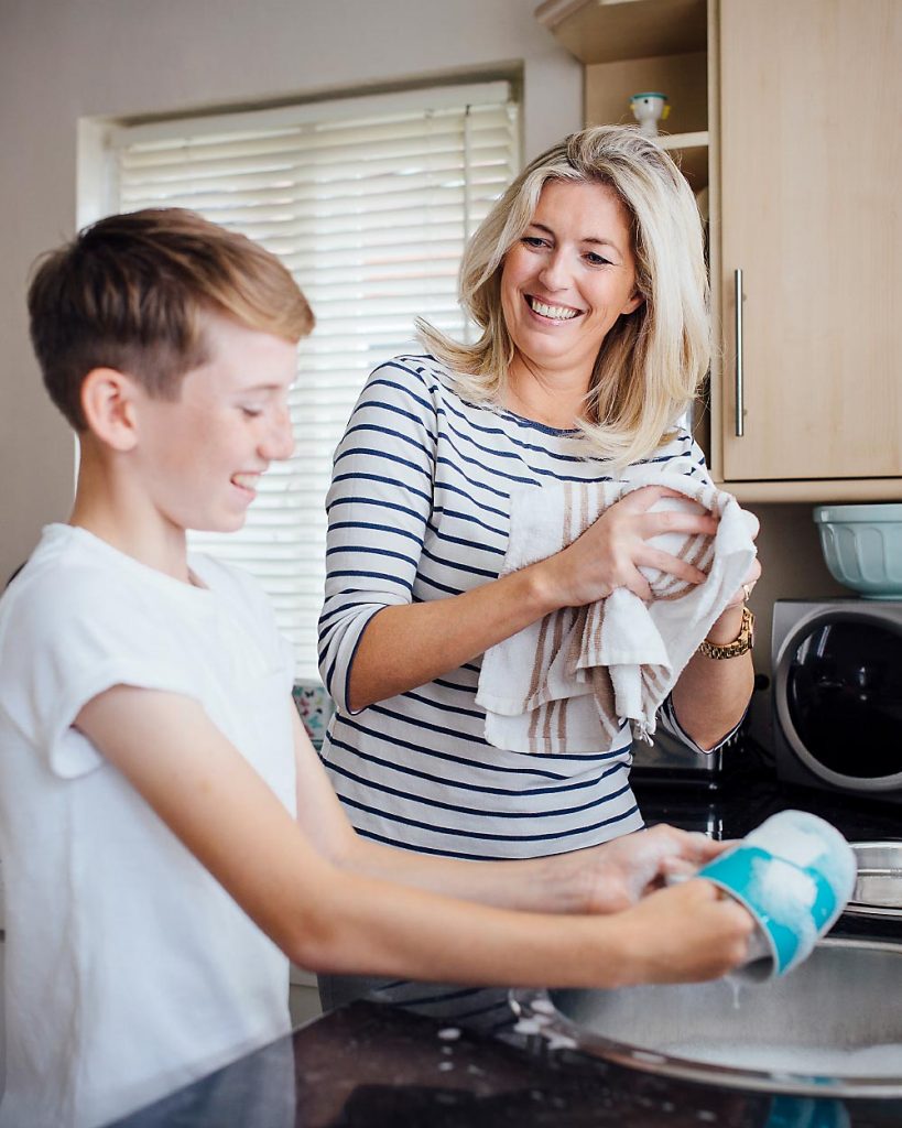 Betreuungskraft macht mit Kind einer Familie die Hilfe benötigt den Abwasch in der Küche am Spülbecken und beide lachen dabei.