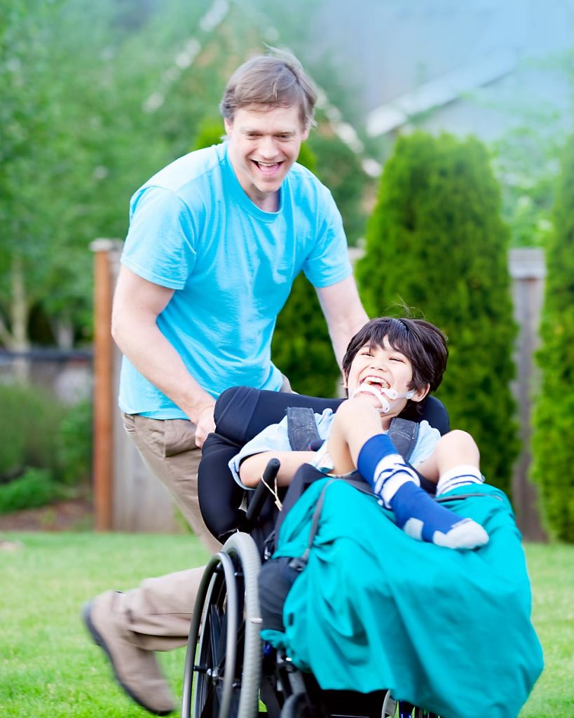Betreuungskraft von Hygge Betreuung schiebt ein Kind, welches im Rollstuhl sitzt und scheibar ein Mensch mit Behinderung ist, durch den grünen Vorgarten. Beide lachen und haben Spaß miteinander.