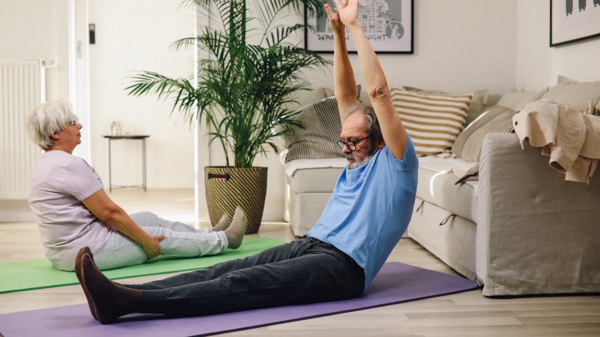 Seniorin und Senior machen auf dem Wohnzimmerfußboden gymnastische Übungen für einen gesunden Rücken.