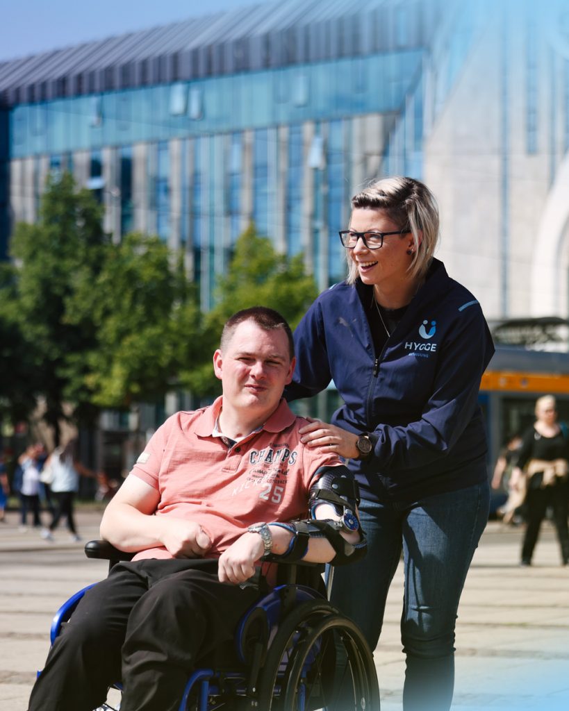 Betreuungskraft von Hygge Betreuung schiebt ein jungen Mann im Rollstuhl, durch die Innenstadt von Leipzig. Beide lachen und genießen den sonnigen Tag.