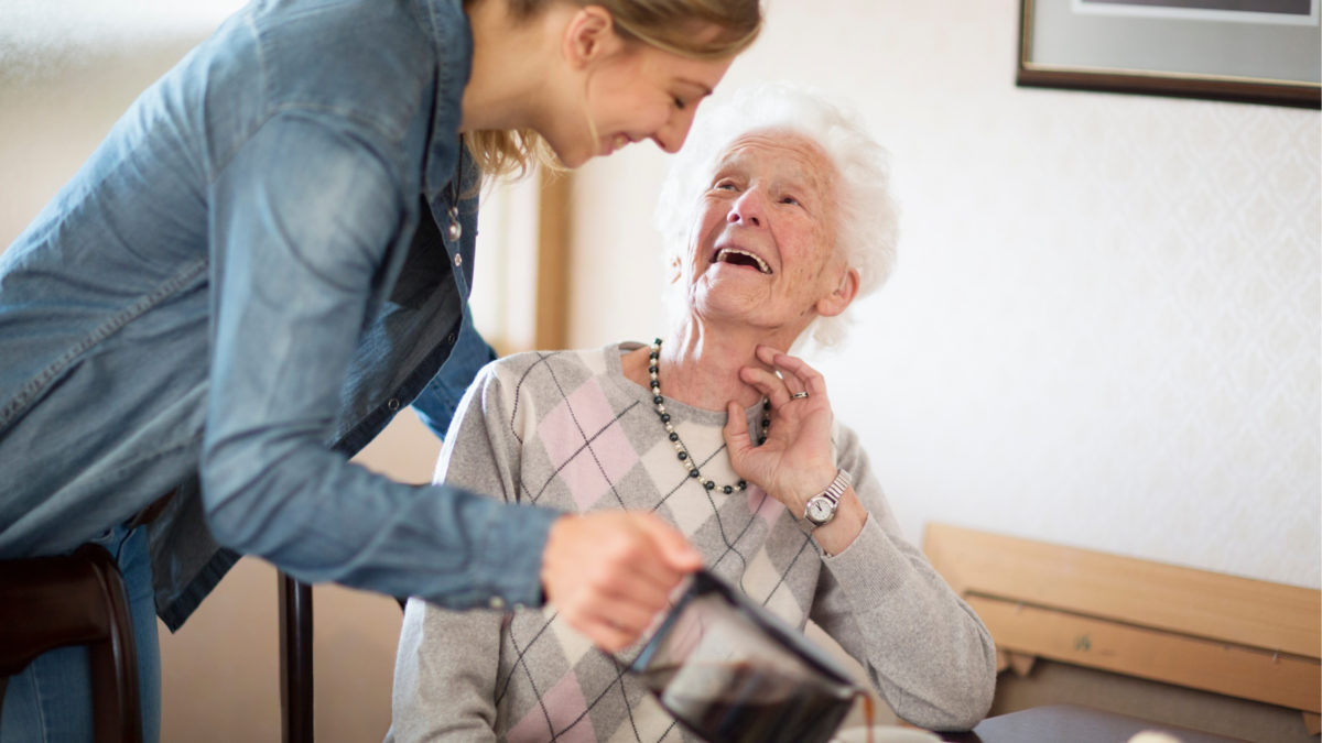 Hygge Betreuung - Tagespflege - junge Frau schenkt Seniorin einen Kaffee ein, beide lächeln.