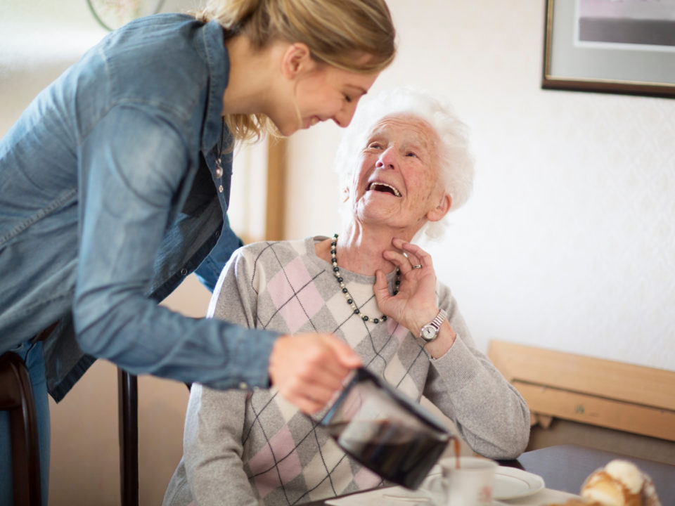 Hygge Betreuung - Tagespflege - junge Frau schenkt Seniorin einen Kaffee ein, beide lächeln.