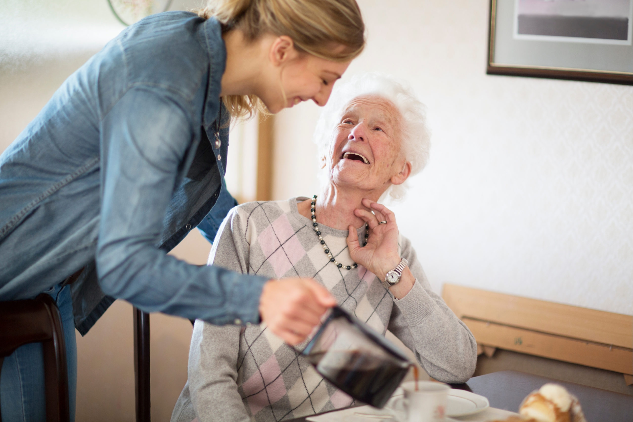 Hygge Betreuung - Tagespflege - junge Frau schenkt Seniorin einen Kaffee ein, beide lächeln.