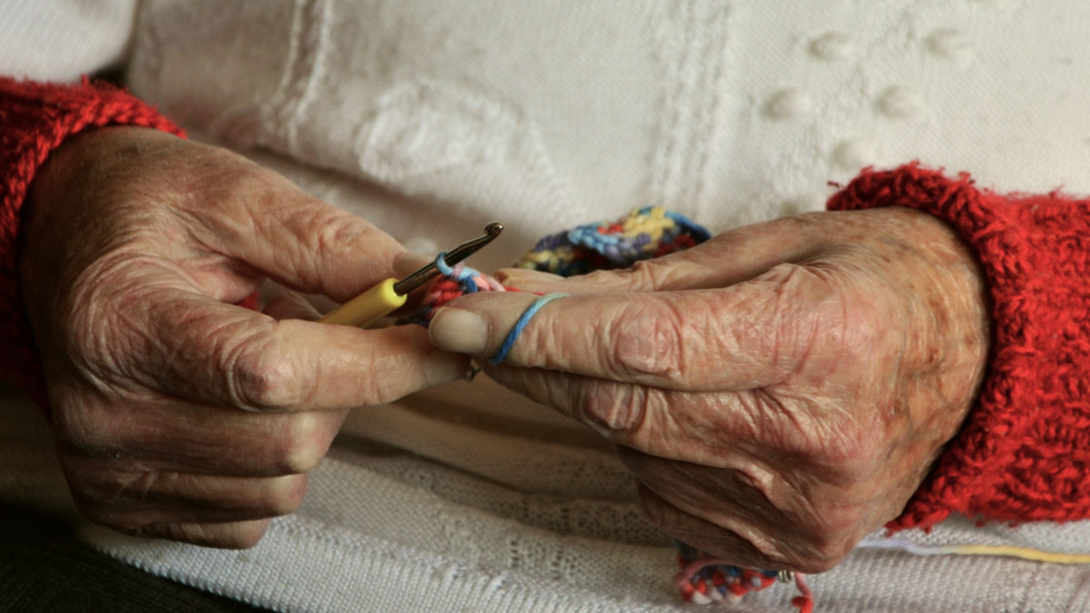 Zwei alte, faltige Hände einer Seniorin in einem weißen Pullover, die beim Häkeln gezeigt werden. Das Bild symbolisiert das Alter und steht im Kontext von Gerontologie.