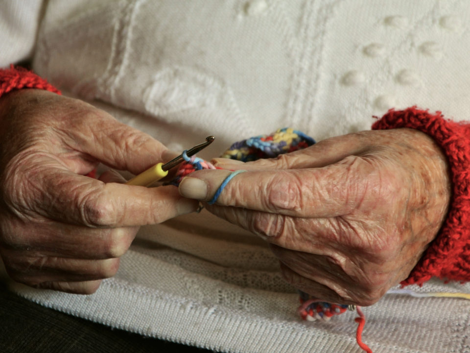 Zwei alte, faltige Hände einer Seniorin in einem weißen Pullover, die beim Häkeln gezeigt werden. Das Bild symbolisiert das Alter und steht im Kontext von Gerontologie.