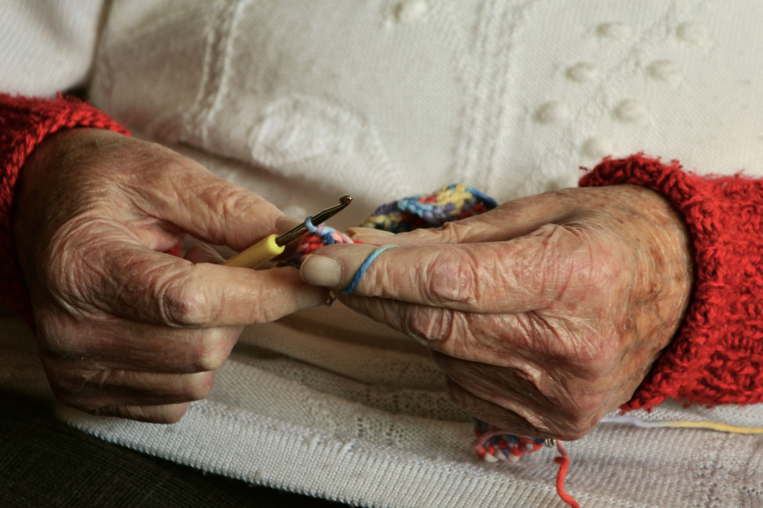 Zwei alte, faltige Hände einer Seniorin in einem weißen Pullover, die beim Häkeln gezeigt werden. Das Bild symbolisiert das Alter und steht im Kontext von Gerontologie.