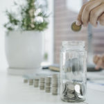 Ein Mann sitzt an einem Tisch und wirft eine 2-Euro-Münze in ein Glas, das mit Geld gefüllt ist. Das Glas steht vor ihm auf dem Tisch und wird mit einer Hand gehalten. Symbolische Darstellung für Beiträge oder Sparen im Kontext der gesetzlichen Pflegeversicherung.