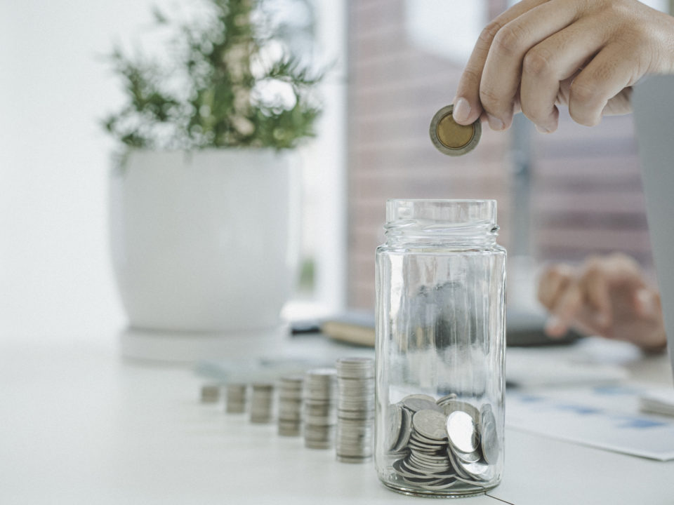 Ein Mann sitzt an einem Tisch und wirft eine 2-Euro-Münze in ein Glas, das mit Geld gefüllt ist. Das Glas steht vor ihm auf dem Tisch und wird mit einer Hand gehalten. Symbolische Darstellung für Beiträge oder Sparen im Kontext der gesetzlichen Pflegeversicherung.